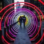 A series of circular lights (about nine or ten feet in diameter) are arrange in a tunnel, with people inside
